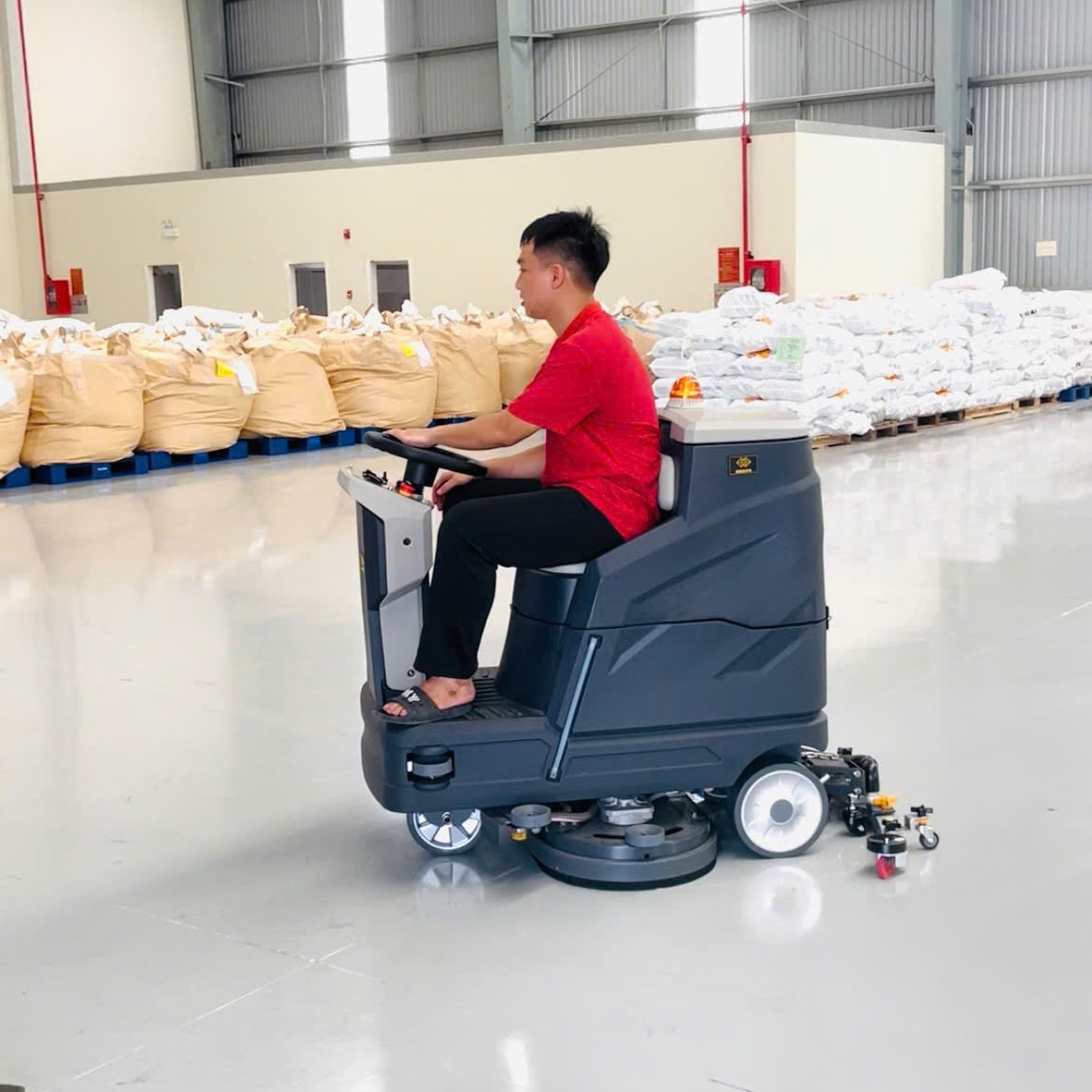 Máy chà sàn ngồi lái Krapo M2913 Ảnh góc rộng của Krapo M2913 trong môi trường nhà kho với giá kệ và hàng hóa.